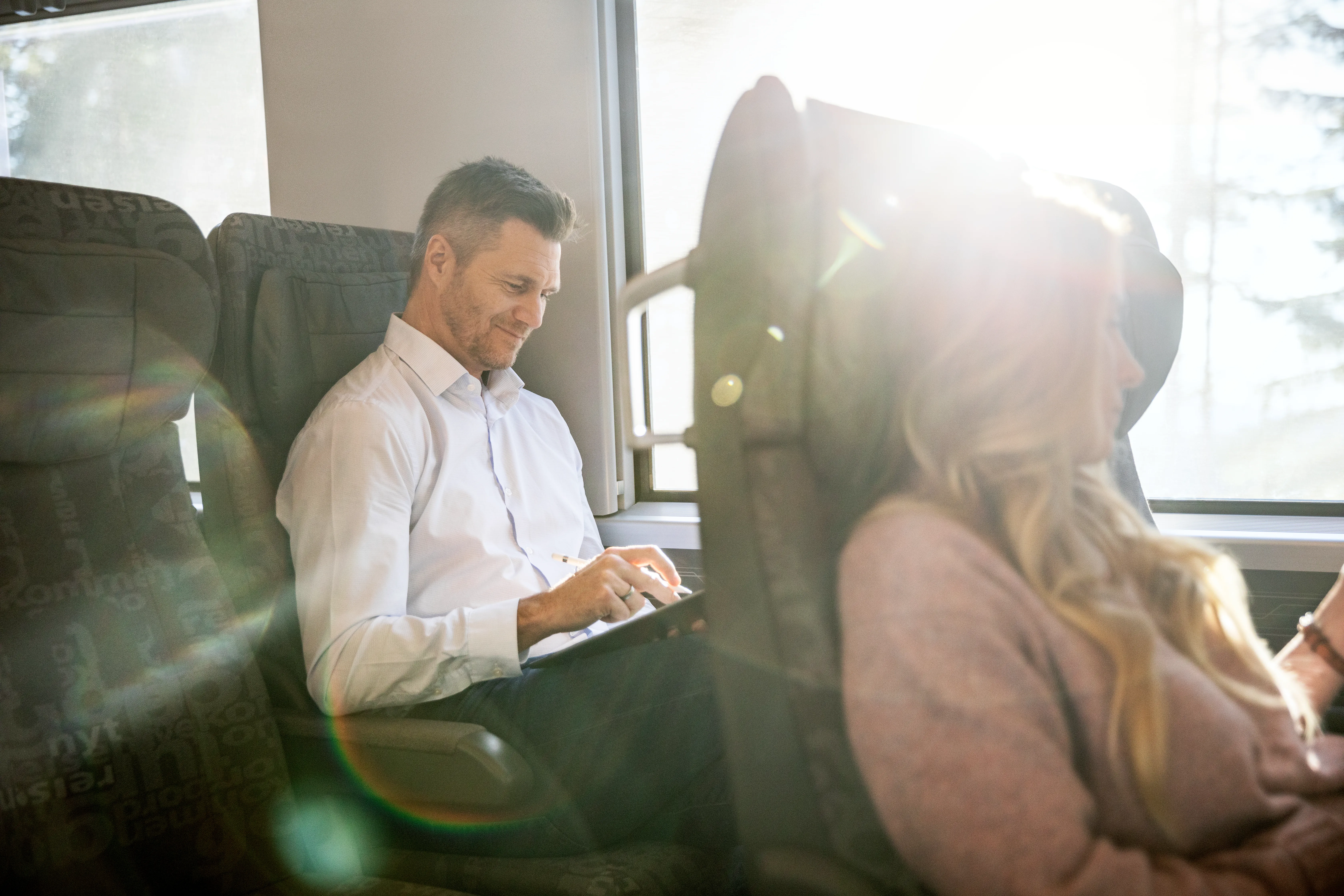 Mann i hvit skjorte sitter med laptop på fanget på toget og smiler