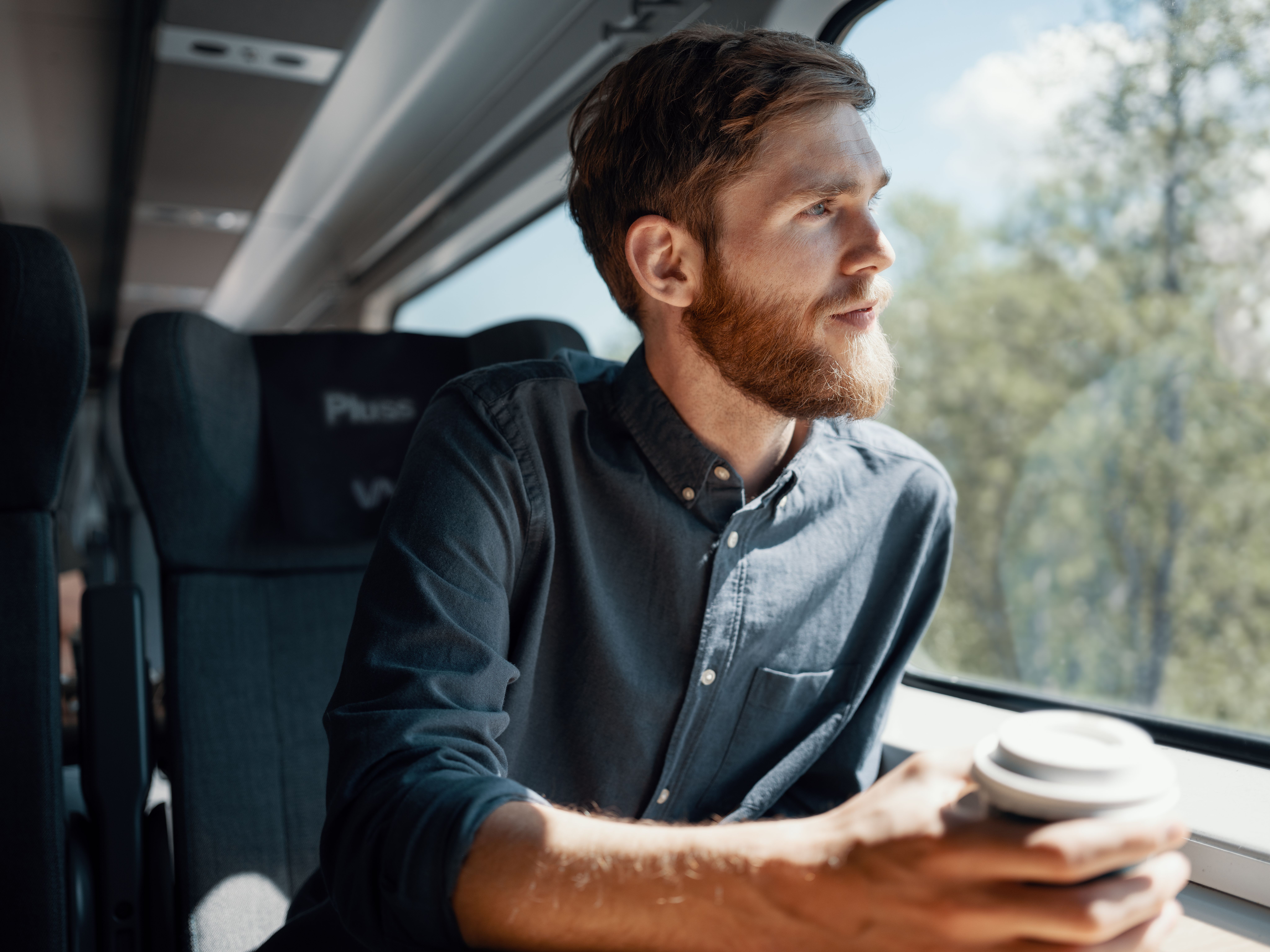 Mann med skjegg og skjorte sitter i Pluss-vogn på Bergensbanen, ser ut vinduet og nyter en kaffe.