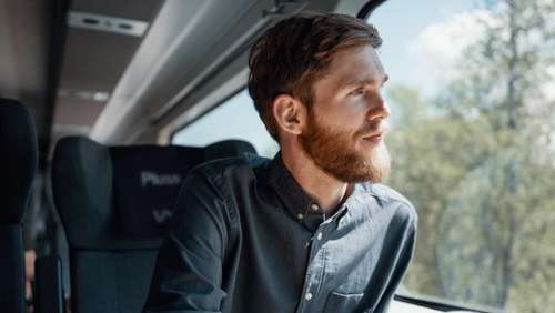 Mann med skjegg og skjorte sitter i Pluss-vogn på Bergensbanen, ser ut vinduet og nyter en kaffe.