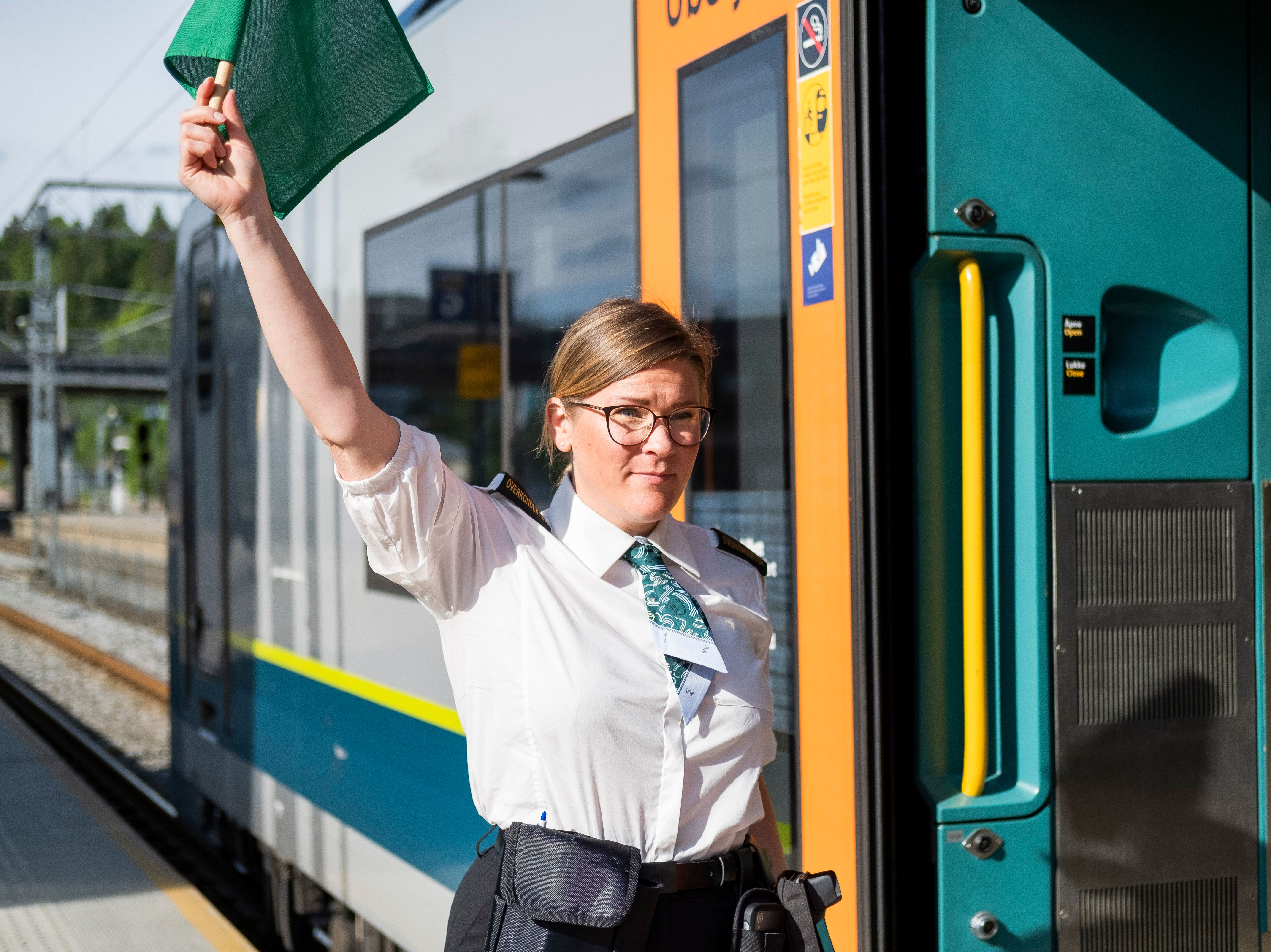 Kvinnelig konduktør med briller står ved toget på stasjonen og vinker med et grønt flagg.