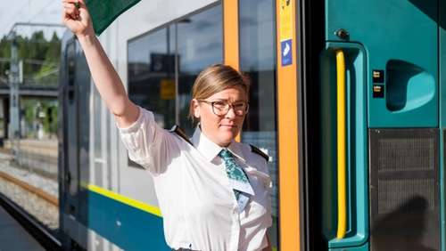 Kvinnelig konduktør med briller står ved toget på stasjonen og vinker med et grønt flagg.