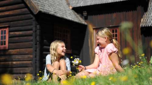 To jenter sitter i gresset utenfor en brun hytte.
