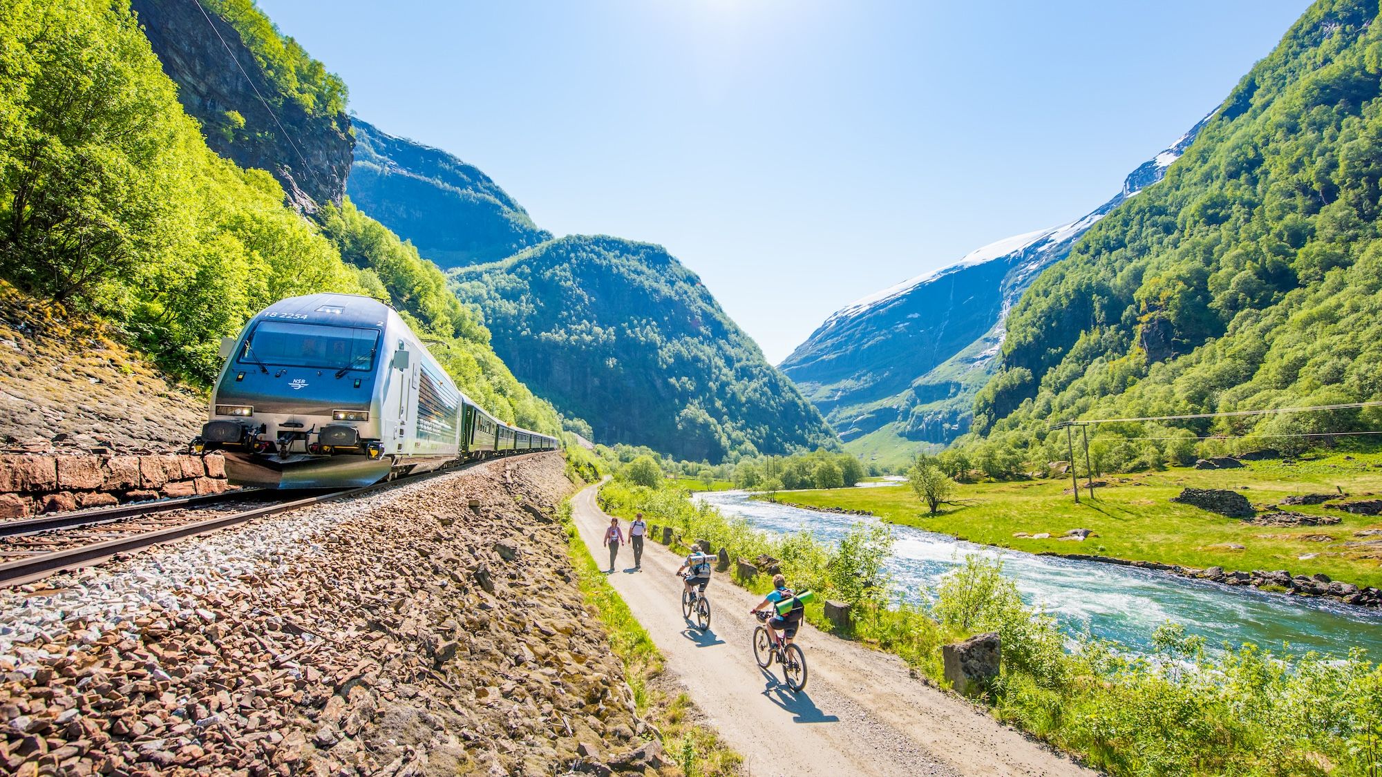Tog som kjører i Flåmsdalen. Personer som sykler og går på en vei ved siden av togsporene.