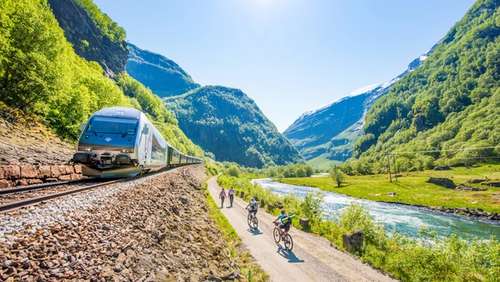 Tog som kjører i Flåmsdalen. Personer som sykler og går på en vei ved siden av togsporene.