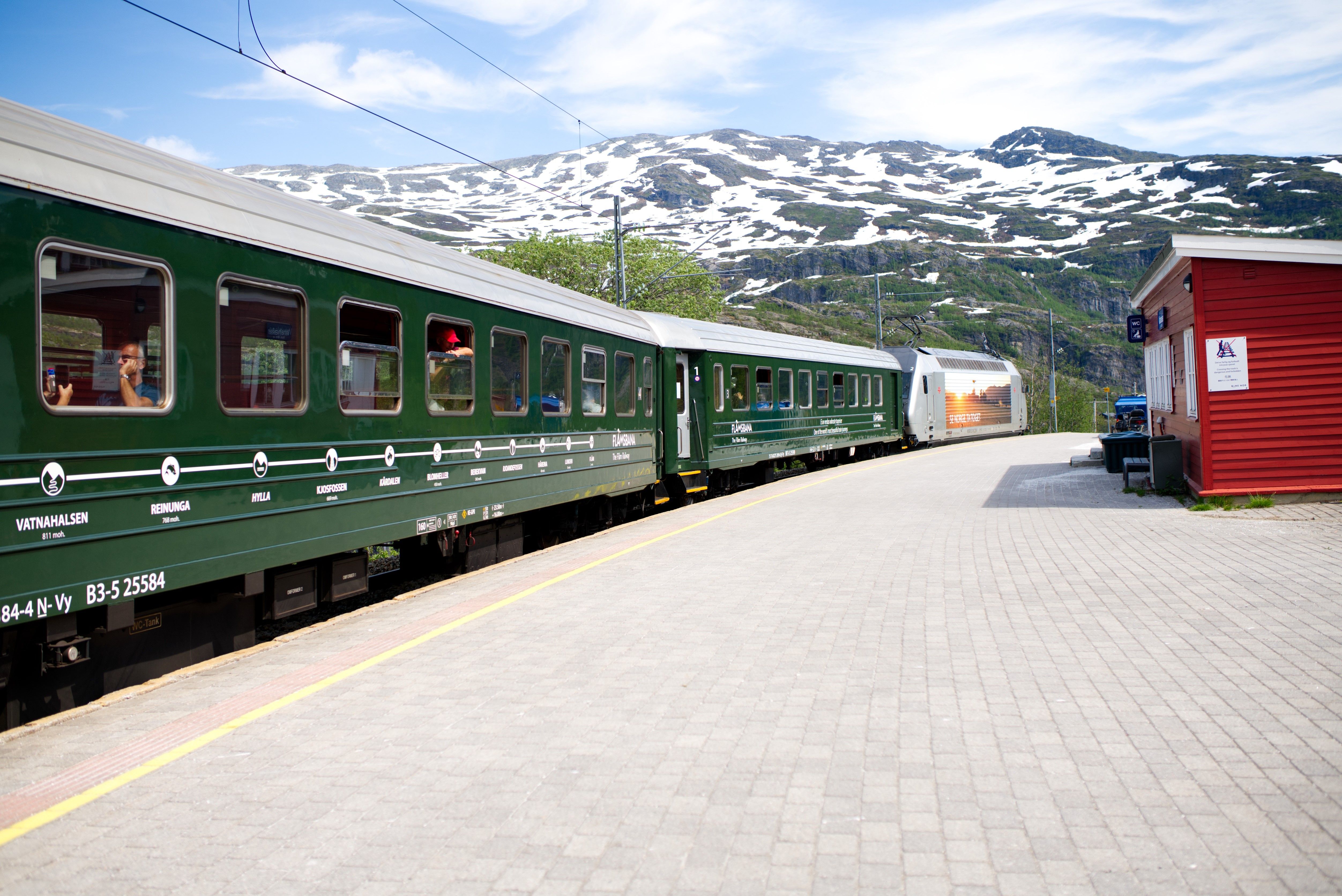 Tog på Flåmsbana på Vatnahalsen stasjon med snøkledde fjell i bakgrunnen