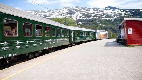 Tog på Flåmsbana på Vatnahalsen stasjon med snøkledde fjell i bakgrunnen