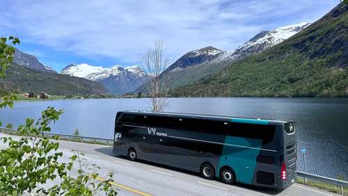 En Vy-buss som står parkert langs fjorden omringet av fjell.