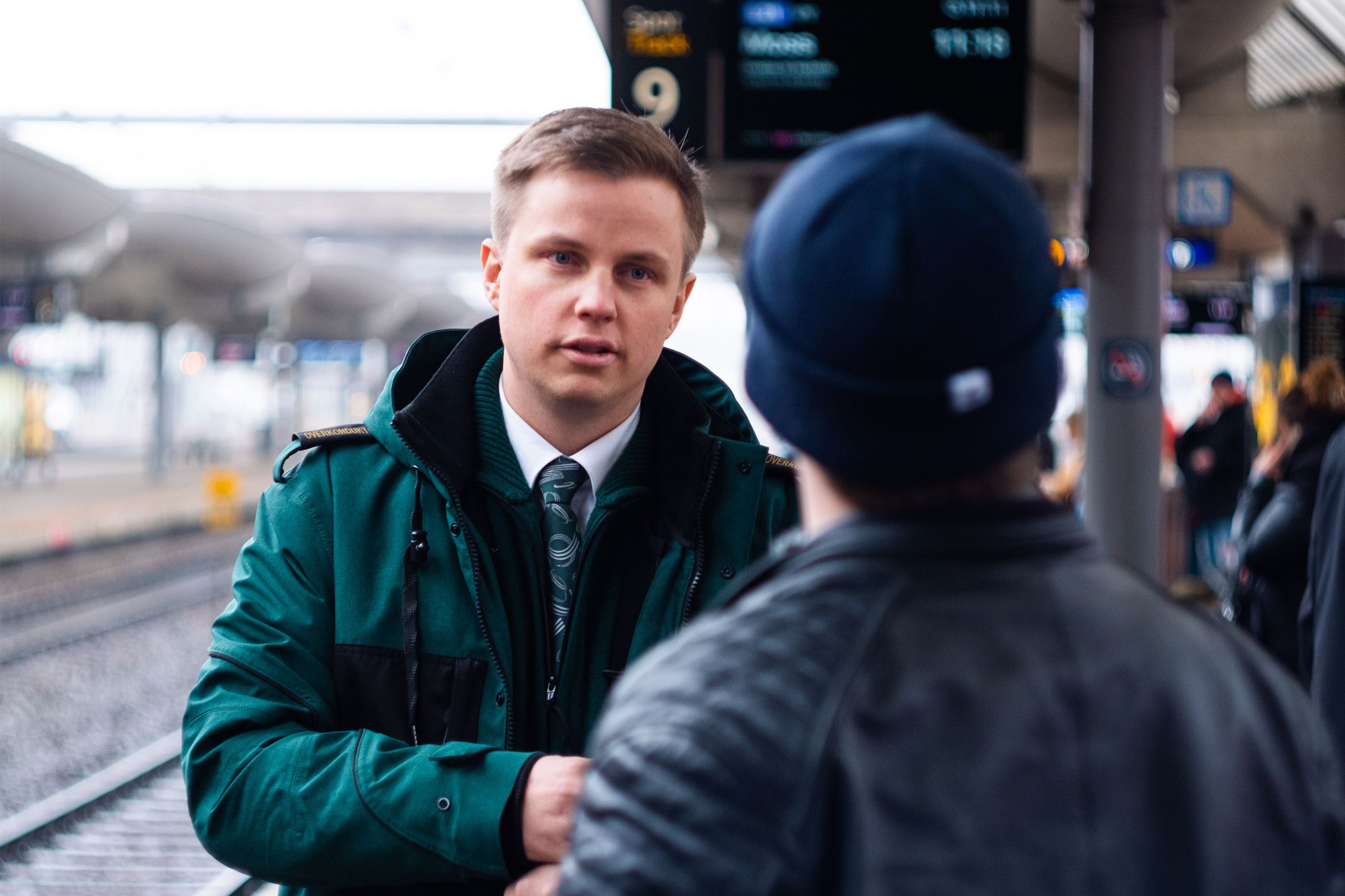 Konduktør Stian iført grønn Vy-jakke, står på en togstasjon og snakker med en passasjer