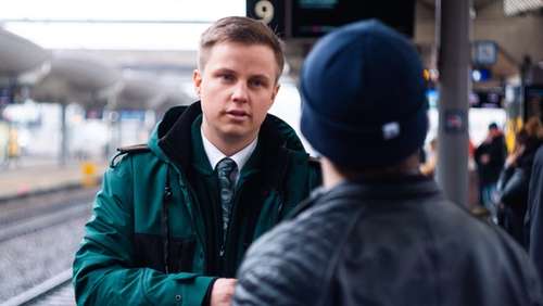 Konduktør Stian iført grønn Vy-jakke, står på en togstasjon og snakker med en passasjer