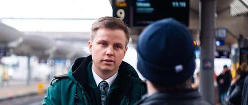 Konduktør Stian iført grønn Vy-jakke, står på en togstasjon og snakker med en passasjer